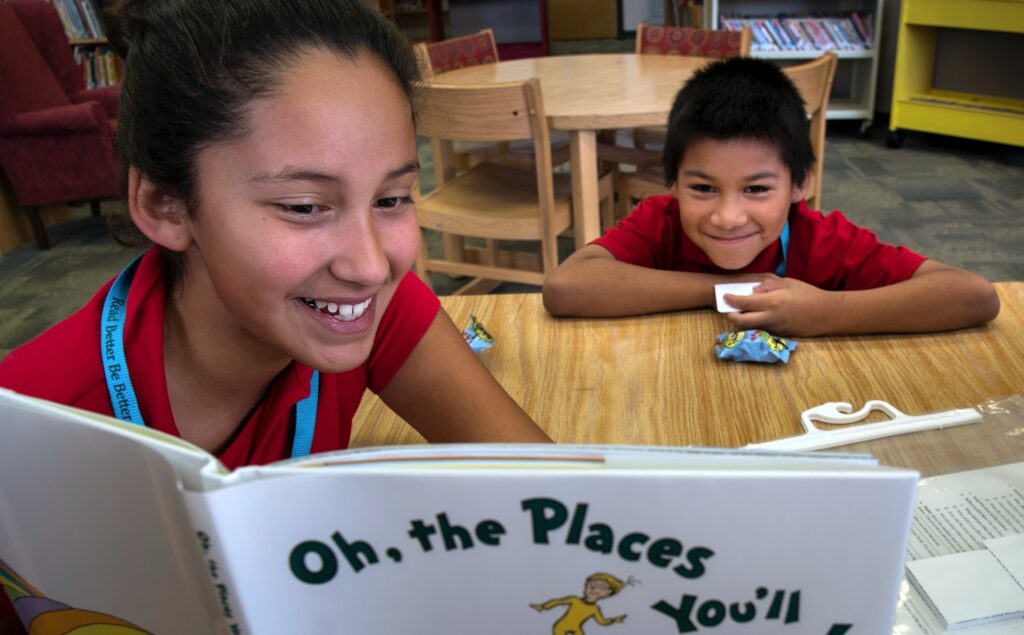 two students are reading "Oh the Places You'll Go" by Dr. Seuss together with smiles on their faces.