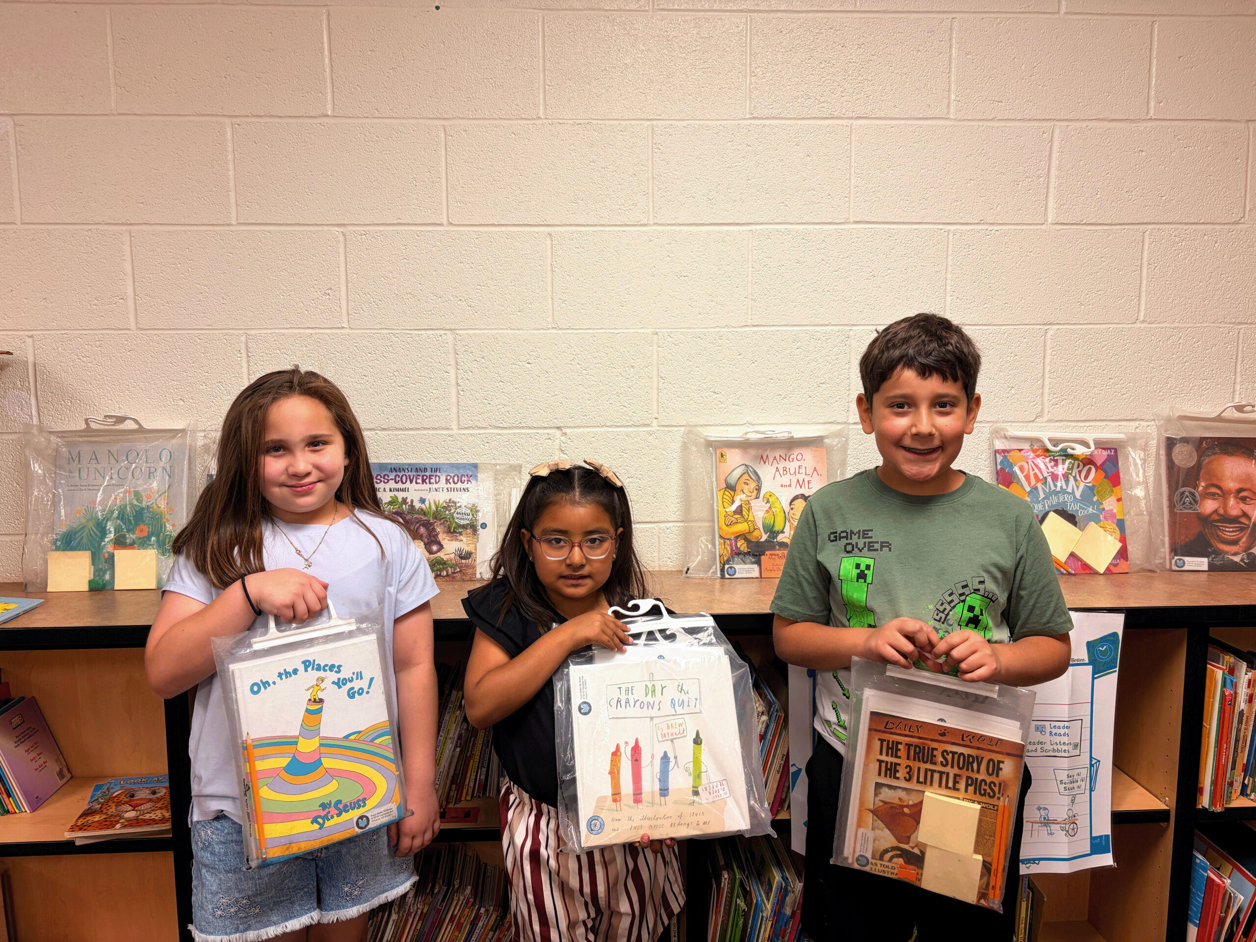 Three elementary students stand smiling in a library in front of a bookshelf, each holding a clear plastic bag containing a children's book and Read Better Be Better curriculum materials